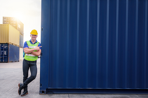 young confident Caucasian man engineer wearing yellow safety helmet standing with blue containers box with copy space from Cargo freight ship for import and export, transport.