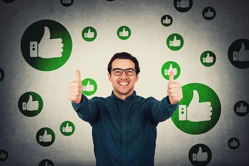 Cheerful businessman shows thumbs up positive gesture, looks to camera isolated over grey wall. Business person wearing eyeglasses, smiling broadly, approval symbol, excellent feedback, multiple likes