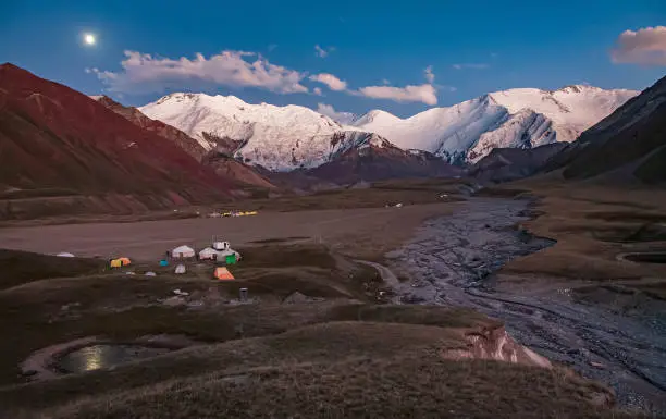 Tent camp with yurts at the foot of the mountains. Scenic landscape of snowy mountain peaks and river. Sunset.