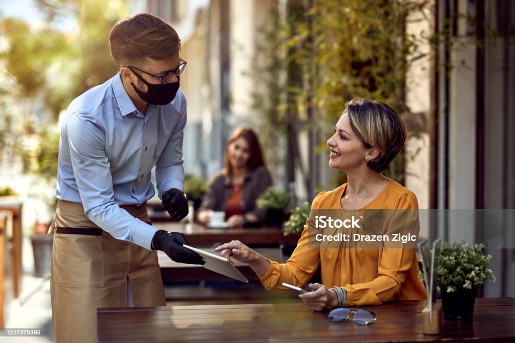 I would like to order this! Happy woman talking to a waiter who is wearing protective face mask while choosing something from a menu on touchpad in a cafe. Restaurant Stock Photo