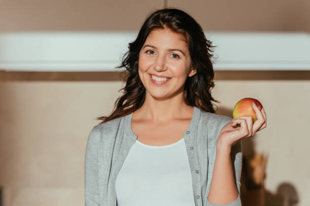 istock Beautiful smiling woman looking at camera while holding fresh apple in kitchen 1227370862