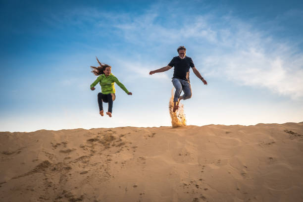 caucasian girl and man jumping over the top of a big sand dune in summer - energia reativa imagens e fotografias de stock