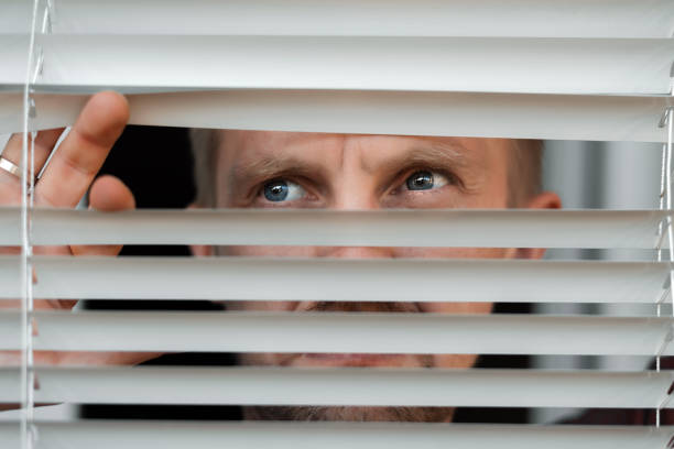 istock Worried man looking looks scared in window peeking out from blinds due to quarantine 1227383104