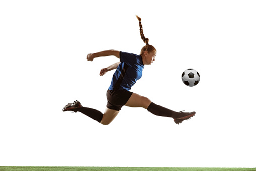 Young female soccer or football player with long hair kicking ball for the goal in flight, jumping high on white studio background. Concept of healthy lifestyle, professional sport, motion, movement.