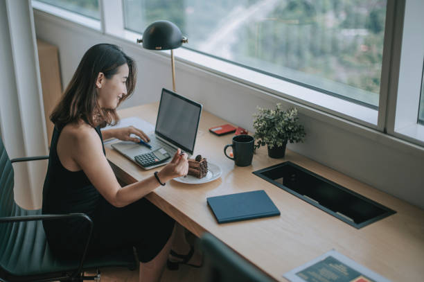 an asian chinese female white collar worker young executive enjoying her chocholate cake while working at her workstation in co-sharing working office in the afternoon with social distancing applied una asiática china trabajadora de cuello blanco joven ejecutiva disfrutando de su pastel de chocholate mientras trabajaba en su estación de trabajo en la oficina de trabajo de co-compartición en la tarde con distanciamiento social aplicado