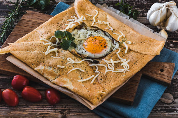 crepe de panqueque francés con huevo frito, hierbas, tomates y queso sobre un listón sobre lino azul sobre una mesa de madera - galette fotografías e imágenes de stock