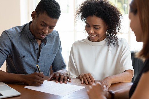 Happy African American family signing contract, legal documents, satisfied clients making deal with Real Estate Agent or broker, smiling couple wife and husband purchasing new house, taking loan or mortgage