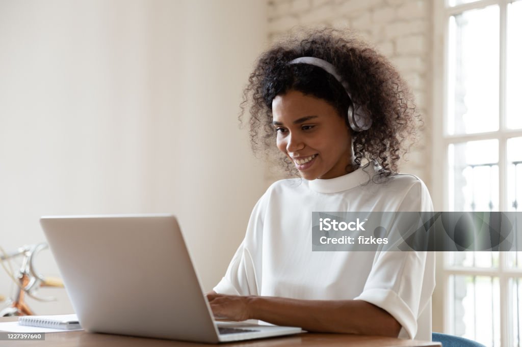 Smiling African American woman wearing headset using laptop in office Smiling African American woman wearing headset using laptop in modern office, looking at screen and typing, listening to music, interpreter working online, learning computer course, lecture Internet Stock Photo