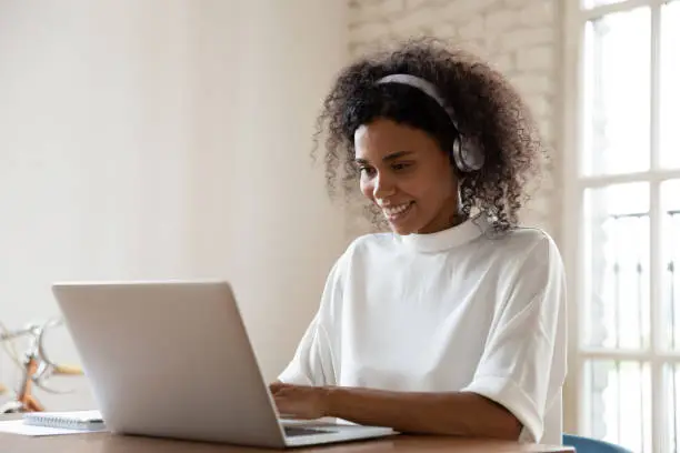 Smiling African American woman wearing headset using laptop in modern office, looking at screen and typing, listening to music, interpreter working online, learning computer course, lecture