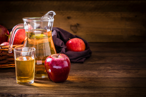 Front view of a crystal jug and a drinking glass full of apple juice on a rustic wooden table. In front of the pitcher is a single red apple and behind is a wicker basket full of multicolored apples. The objects are at the left side of the image leaving a useful copy space at the right  on the table. Low key DSLR photo taken with Canon EOS 6D Mark II and Canon EF 24-105mm f / 4