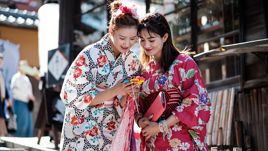 Two smiling ladies dressed as geisha with beautiful back light looking at their smart phones in the middle of the street, Kyoto Japan.