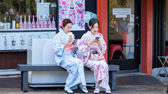Traditionally dressed ladies sitting on a bench in front of a liquor store with similar colors, both looking at their smart phones. one of them eating ice cream, Kyoto, Japan.