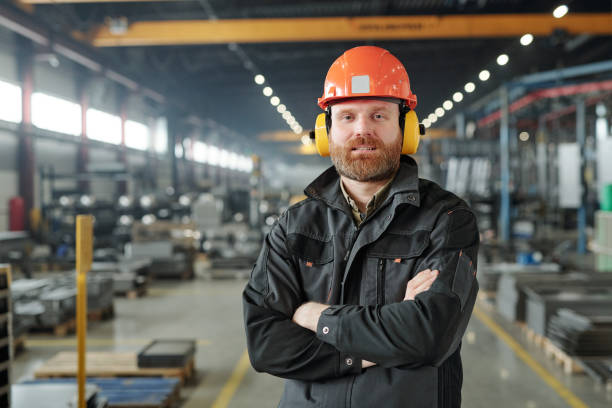 Bearded factory worker in ear protectors Portrait of content bearded factory worker in ear protectors and hardhat standing with crossed arms in contemporary shop of industrial plant ear protectors stock pictures, royalty-free photos & images