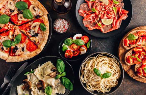 full table of italian meals on plates pizza, pasta, ravioli, carpaccio. caprese salad and tomato bruschetta on black background. - italian culture imagens e fotografias de stock