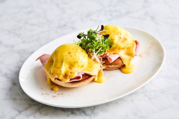 eggs Benedict with arugula, bacon and hollandaise sauce on gray wooden background. Eggs Benedict on a white plate, side view. Side view on eggs Benedict with arugula, bacon and hollandaise sauce on gray wooden background. hollandaise sauce stock pictures, royalty-free photos & images