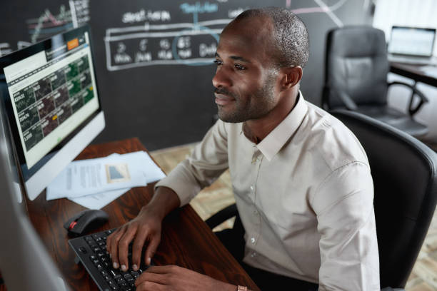 Earn more and count more. Cheerful african male trader sitting by desk and studying analytical reports using pc in the office. Cheerful african male trader sitting by desk and studying analytical reports using pc in the office. Stock trading, people concept. Horizontal shot stock certificate photos stock pictures, royalty-free photos & images