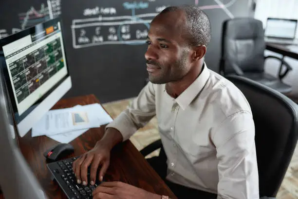 Photo of Earn more and count more. Cheerful african male trader sitting by desk and studying analytical reports using pc in the office.