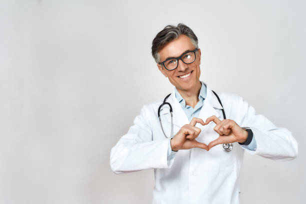 medico maschio maturo felice che indossa uniforme e occhiali, con uno stetoscopio intorno al collo che mostra il segno del cuore e sorride, in piedi su sfondo grigio - doctor male doctor mature men portrait foto e immagini stock