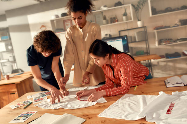istock The total printing arrangement. Creative people trying on stickers with text, while discussing logo and design of T-shirt. Young man and women working at custom T-shirt, clothing printing company 1227433627