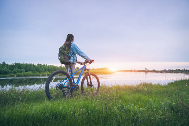 春の日没時に川の近くの丘の上に自転車を持つ女性。夏のマウンテンバイク、未舗装道路、緑の草、水に乗ってバックパックを持つスポーティな女の子と風景。スポーツと旅行。サイクル - landscape hill green grass ストックフォトと画像