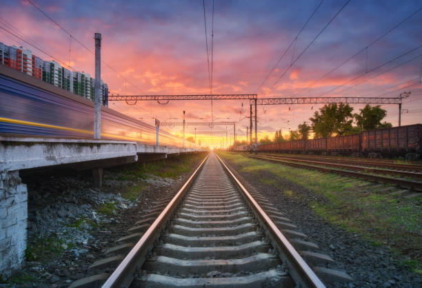 Railway station with blurred high speed train and freight train at colorful sunset. Railroad in summer. Industrial landscape with moving train, railway platform, sky with pink clouds. Transportation