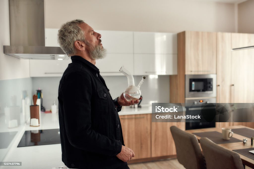 Feel everything. Bearded midle-aged man holding a bong or glass water pipe while smoking marijuana, standing in the kitchen. Cannabis and weed legalization concept Bearded midle-aged man holding a bong or glass water pipe while smoking marijuana, standing in the kitchen. Cannabis and weed legalization concept. Side view. Horizontal shot Bong Stock Photo