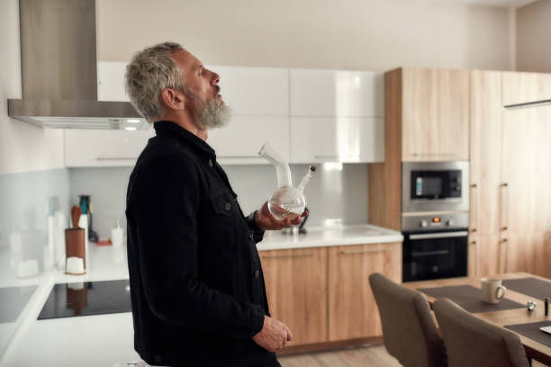 Feel everything. Bearded midle-aged man holding a bong or glass water pipe while smoking marijuana, standing in the kitchen. Cannabis and weed legalization concept Bearded midle-aged man holding a bong or glass water pipe while smoking marijuana, standing in the kitchen. Cannabis and weed legalization concept. Side view. Horizontal shot bong stock pictures, royalty-free photos & images