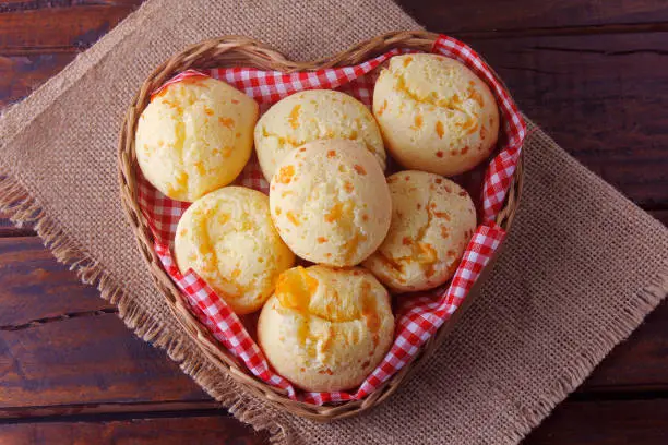 homemade cheese bread, traditional Brazilian snack, in a heart-shaped basket on a rustic kitchen table. Top view