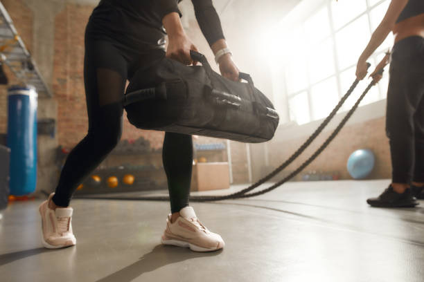 vaincre votre faiblesse. coup recadré de femme sportive soulevant un sac de sable lourd tout en ayant l’entraînement à la gymnastique industrielle. formation de groupe, concept de travail d’équipe - gym bag photos et images de collection