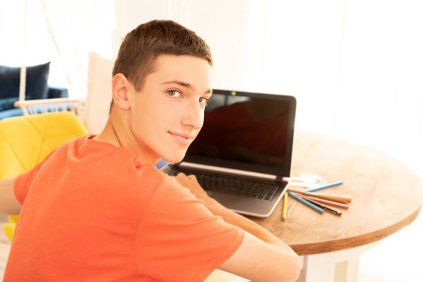 Teenage boy doing homework using computer sitting by desk in room alone.