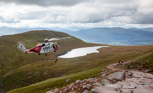 Helicopter came to fly down a tourist which collapsed during descent from Ben Nevis. On the picture we see rescuer hanging on the rope beneath the helicopter.  In the background there`s pond, valley and other mountains.