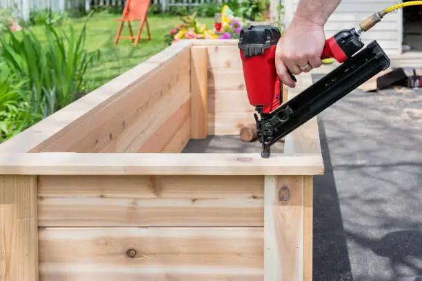 Closeup of a man using a pneumatic nail gun to finish the trim on cedar garden planters with sawdust flying