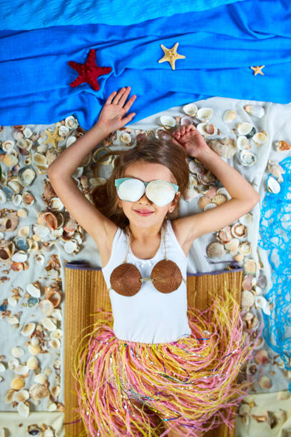 Little girl imitates Havaii vacation at home quarantine. Coronavirus home summer activity, funny, happy, crazy. The child is lying on a fictional beach near the sea or ocean.