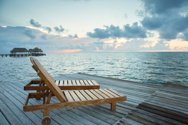 Wooden lounge chairs at the end of the dock with fluffy clouds and pink setting sun. Calm ocean in background stock photo