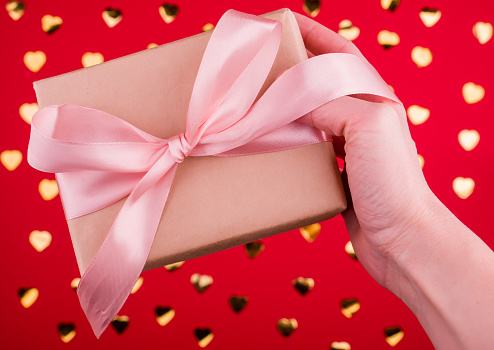 A beautiful gift box with satin ribbon in hand on a red background with golden hearts