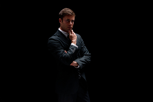 Waist up of aged 20-29 years old with brown hair caucasian male businessman in front of black background wearing businesswear who is contemplating