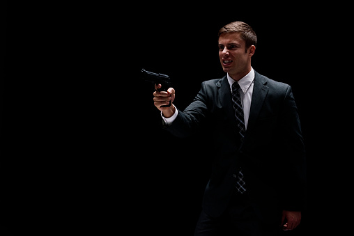 Front view of aged 20-29 years old with short hair caucasian young male business person in front of black background wearing businesswear who is agressive who is shooting a weapon and holding weapon and using handgun
