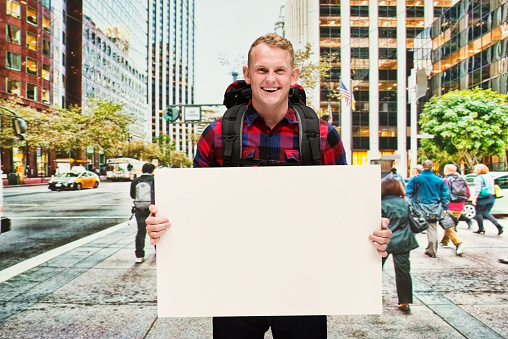 Front view of with redhead caucasian male who is outdoors wearing plaid shirt who is laughing who is showing with hand and holding sign with copy space