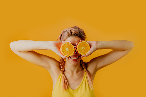 Photo of a playful young woman holding orange halves in front of her eyes; studio shot, isolated on an orange background.