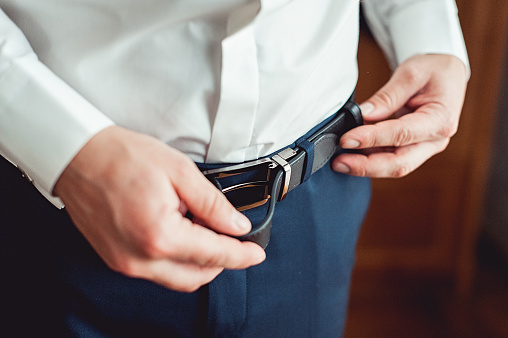 Groom in blue pants and a white shirt fastens his leather belt. grooms mourning. Wedding details. Concept and composition.