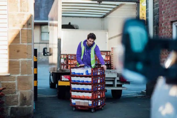 A mid adult Caucasian delivery man pushing bread pallets outdoors, after taking them out of a delivery truck. He is an essential worker during the COVID-19 pandemic.