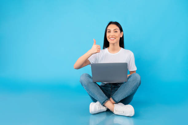 Happy teenager isolated over blue studio background Educational Concept. Excited girl sitting on floor with laptop, showing thumb up gesture, blue studio wall ok sign photos stock pictures, royalty-free photos & images