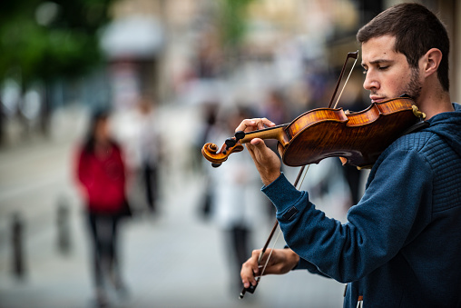 Sofia, Bulgaria - May 26 2020: A young musician is playing the violin on a street in the Sofia city center while people in protective masks are passing around him