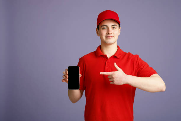 istock Cute courier in red uniform points finger at smartphone with blank screen, isolated gray background 1227504412