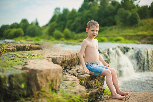 A boy in shorts sits on a stone near a waterfall