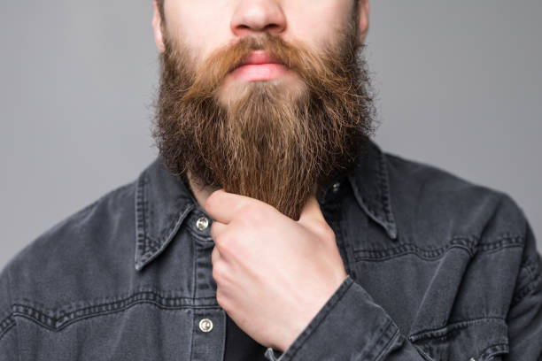 Touching his perfect beard. Close-up of young bearded man touching his beard while standing against grey background