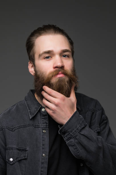 istock Handsome young bearded man is keeping hand on beard and looking away on a gray background 1227514518
