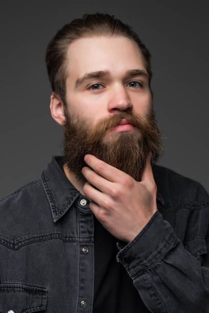 istock Handsome young bearded man is keeping hand on beard and looking away on a gray background 1227516038