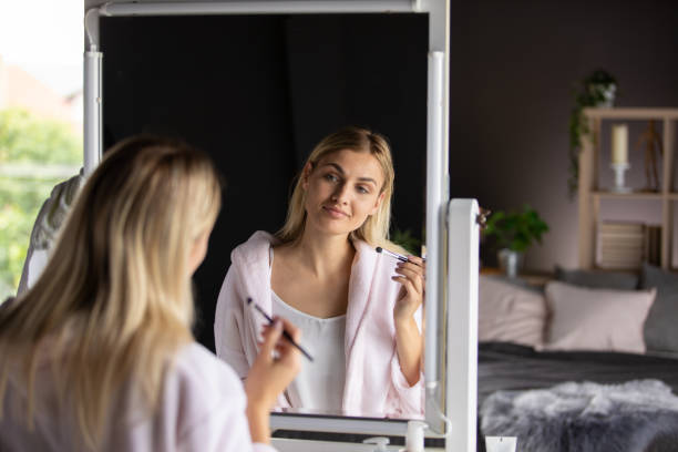 istock Woman in bathrobe is looking in the mirror, holding make-up brush 1227520355