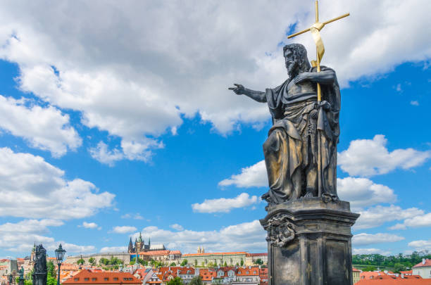 Saint John the Baptist statue on Charles Bridge Karluv Most over Vltava river with Prague Castle, St. Vitus Cathedral in Hradcany district, blue sky white clouds background, Bohemia, Czech Republic stock photo